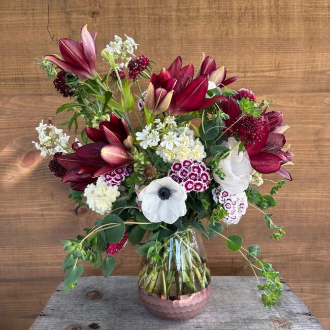 A glass vase holds a mixed bouquet of red lilies, white anemones, small white and burgundy flowers, and green foliage on a wooden table with a wood panel background.