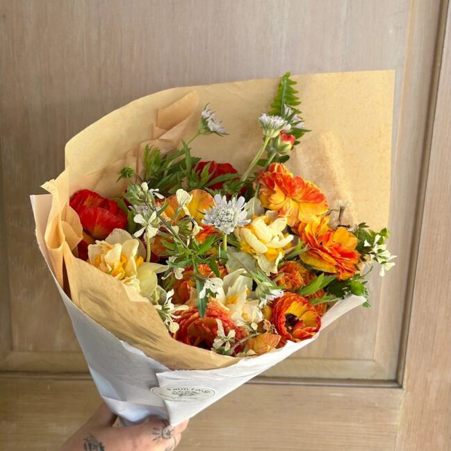 A hand holds a bouquet of assorted flowers in yellow, orange, red, and white, wrapped in brown and white paper, against a wooden background.