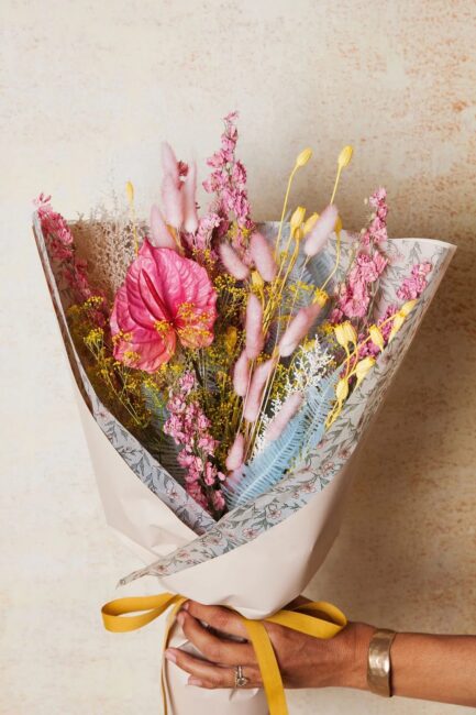 A hand holds a bouquet of assorted dried and fresh flowers in pink, yellow, and blue tones, wrapped in patterned paper and tied with a yellow ribbon.