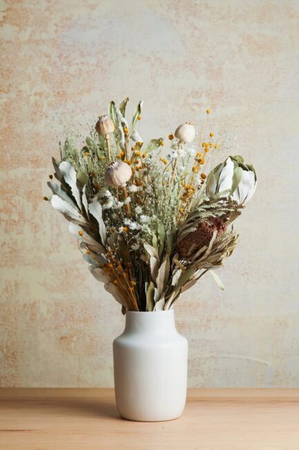 A white ceramic vase holds a mixed arrangement of dried flowers and foliage, displayed on a light wooden surface against a beige textured wall.