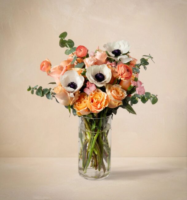 A glass vase filled with a bouquet of peach and white flowers, including roses and anemones, with green foliage, set against a beige background.