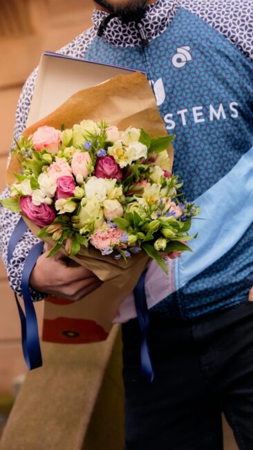 A person wearing a blue-patterned jacket holds a bouquet of mixed flowers wrapped in brown paper and tied with blue ribbon.