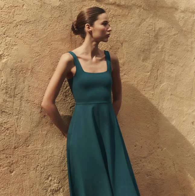 A woman in a teal sleeveless dress stands against a textured beige wall, looking to her left with her hair pulled back in a neat bun.
