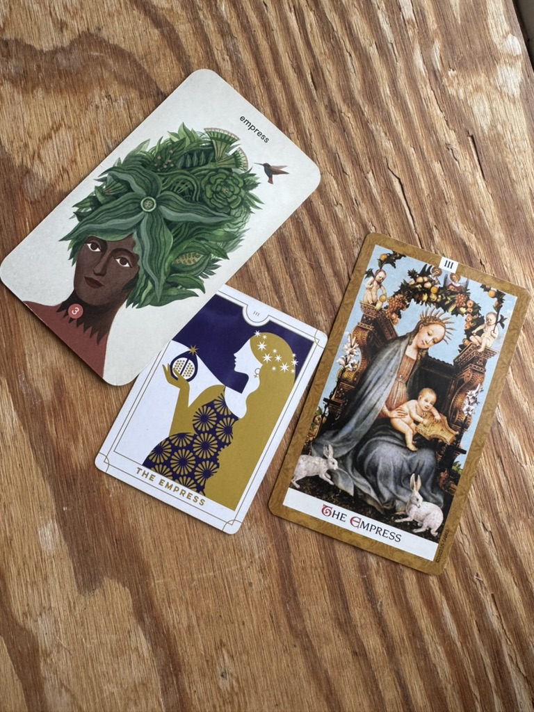 Three Empress tarot cards from different decks are displayed on a wooden surface, each featuring unique artistic interpretations of the Empress archetype.