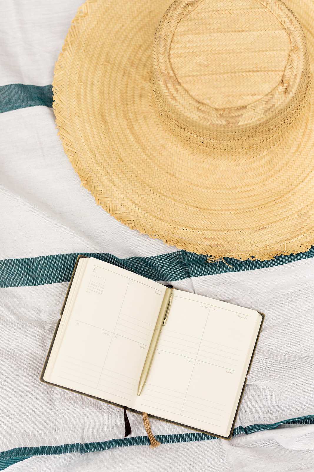 A straw hat and an open planner with a pen lie on a white and green striped fabric, ready to jot down notes for the upcoming July sales.