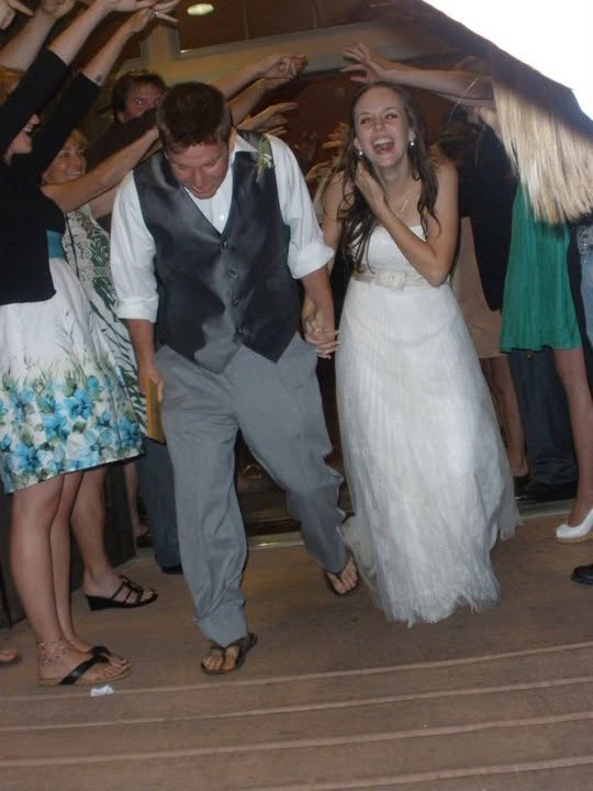 A newlywed couple walks hand in hand through a crowd of cheering guests, who are forming an arch with their arms in celebration.