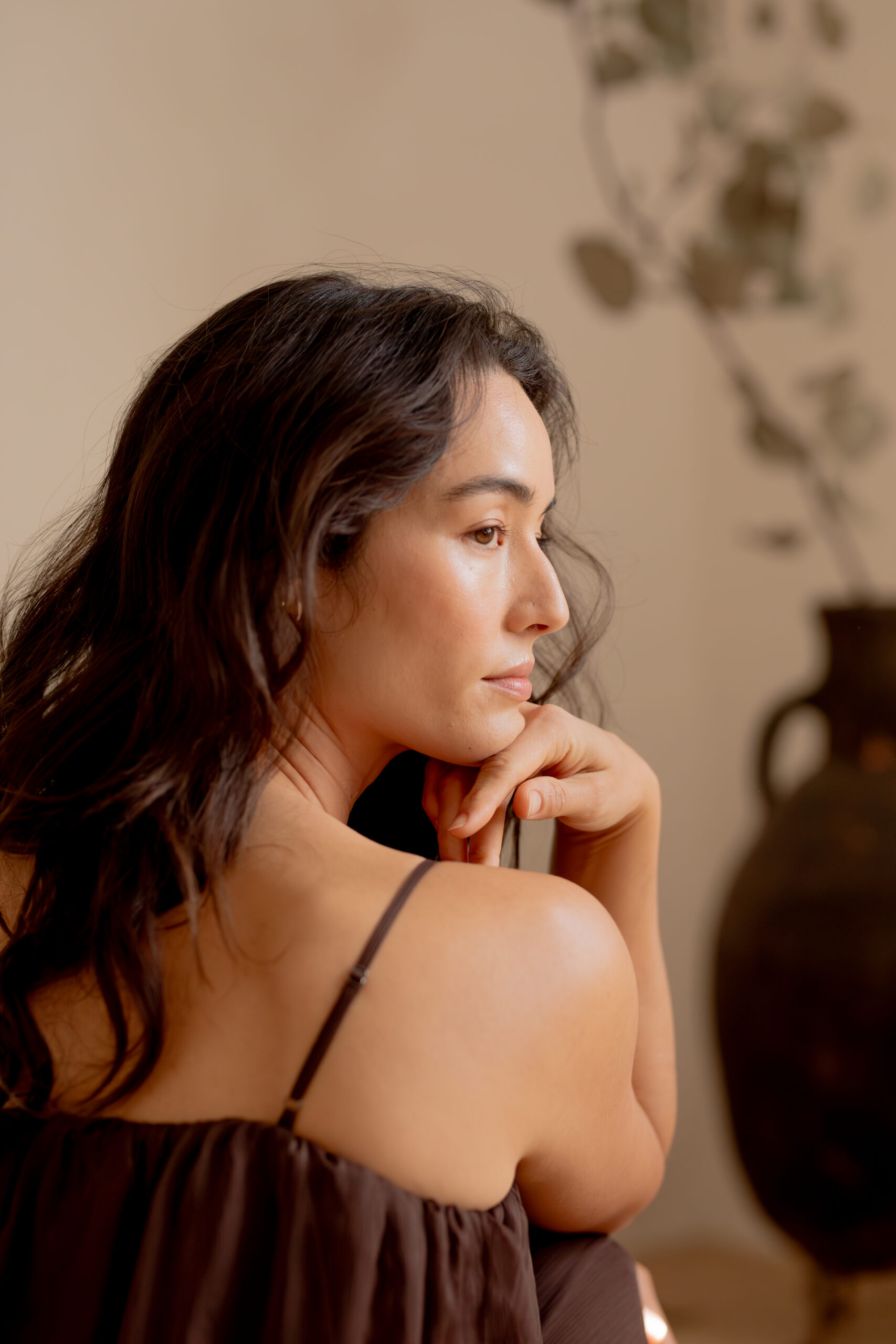 Woman with long dark hair sits in profile with her chin resting on her hand, wearing a dark sleeveless top; a blurred vase is in the background.