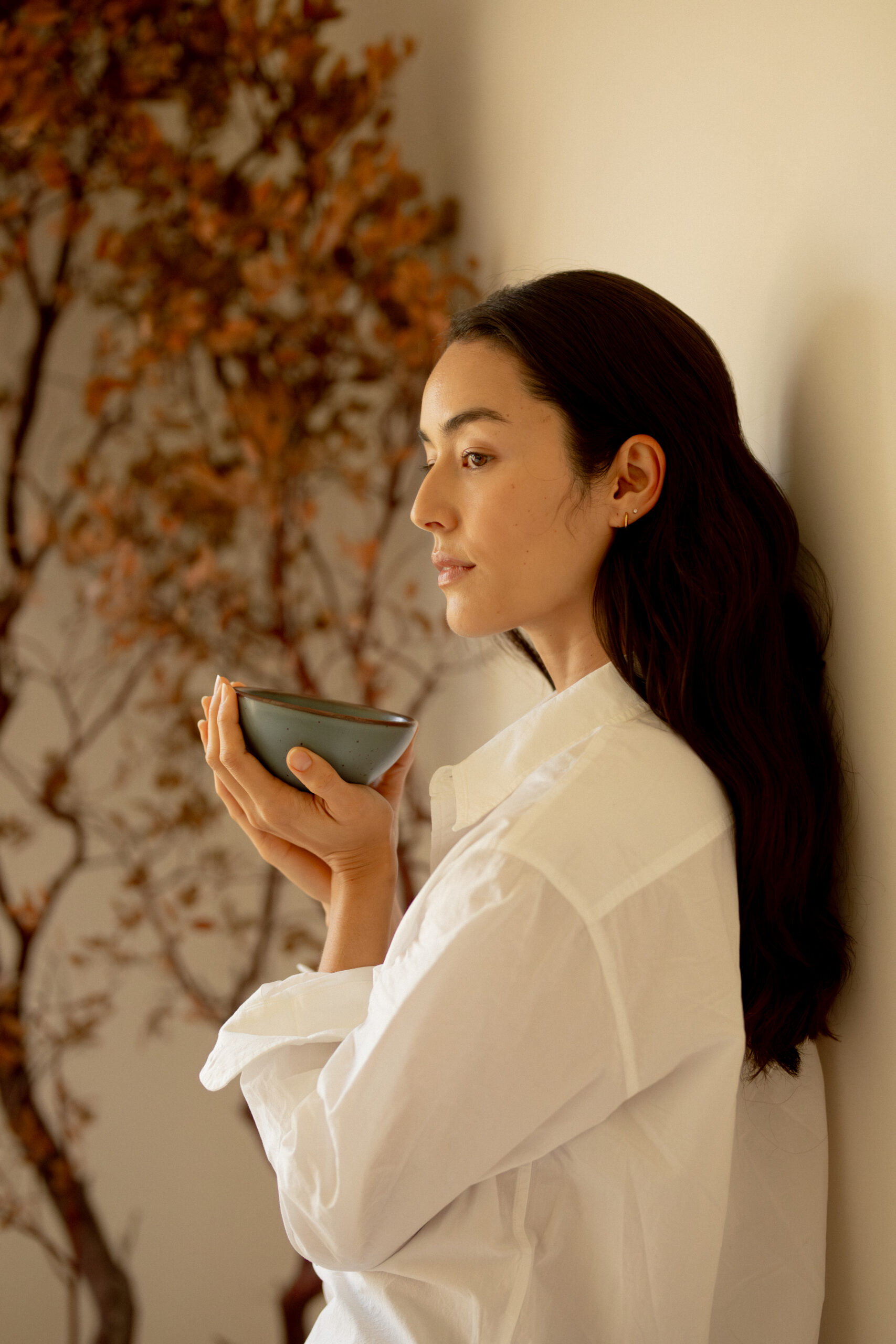 Woman in a white shirt holds a ceramic bowl near her face while standing indoors beside a plant with brown leaves.
