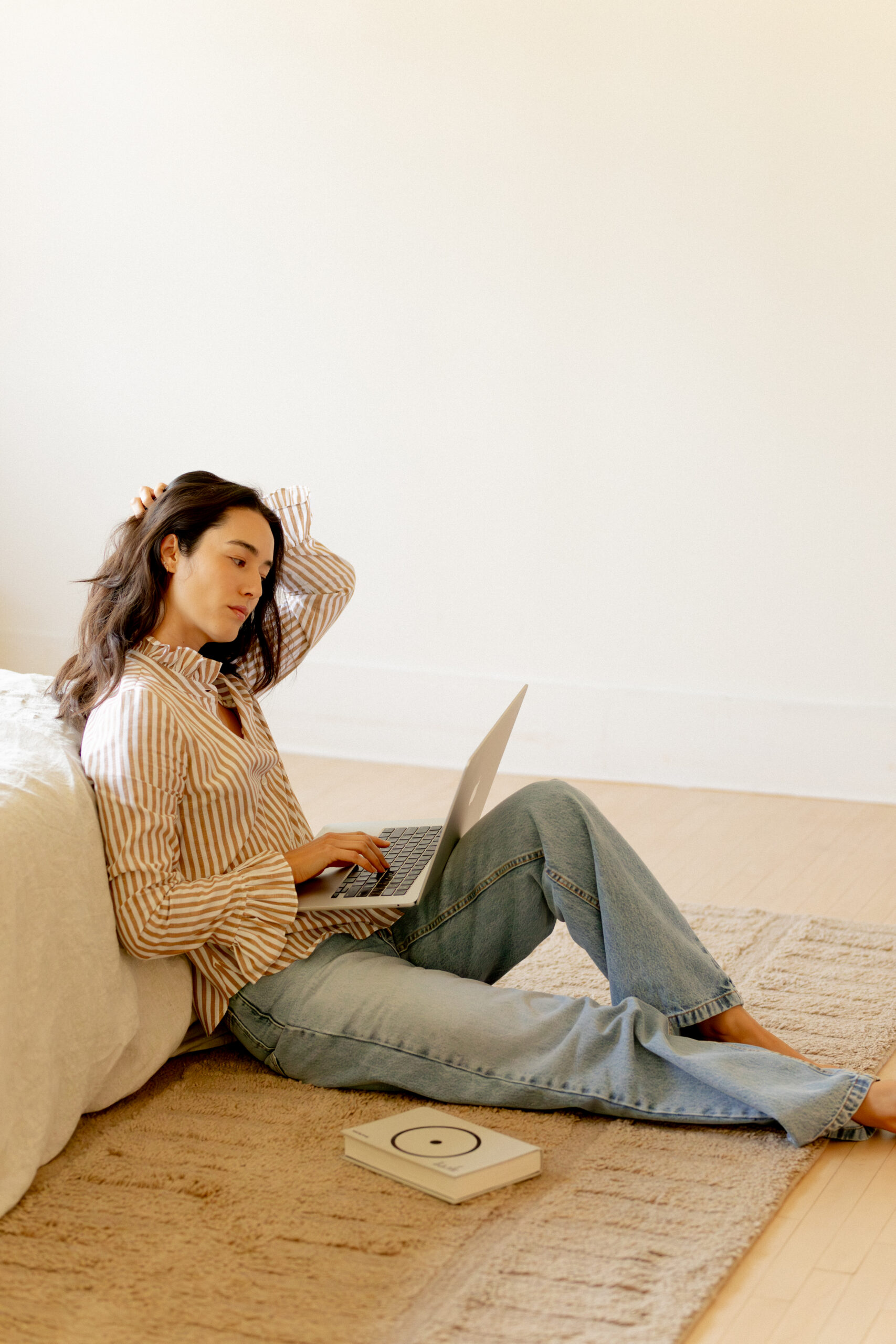 A person sits on a rug next to a bed, using a laptop. A closed book or box lies on the floor nearby. The room is minimal and brightly lit.