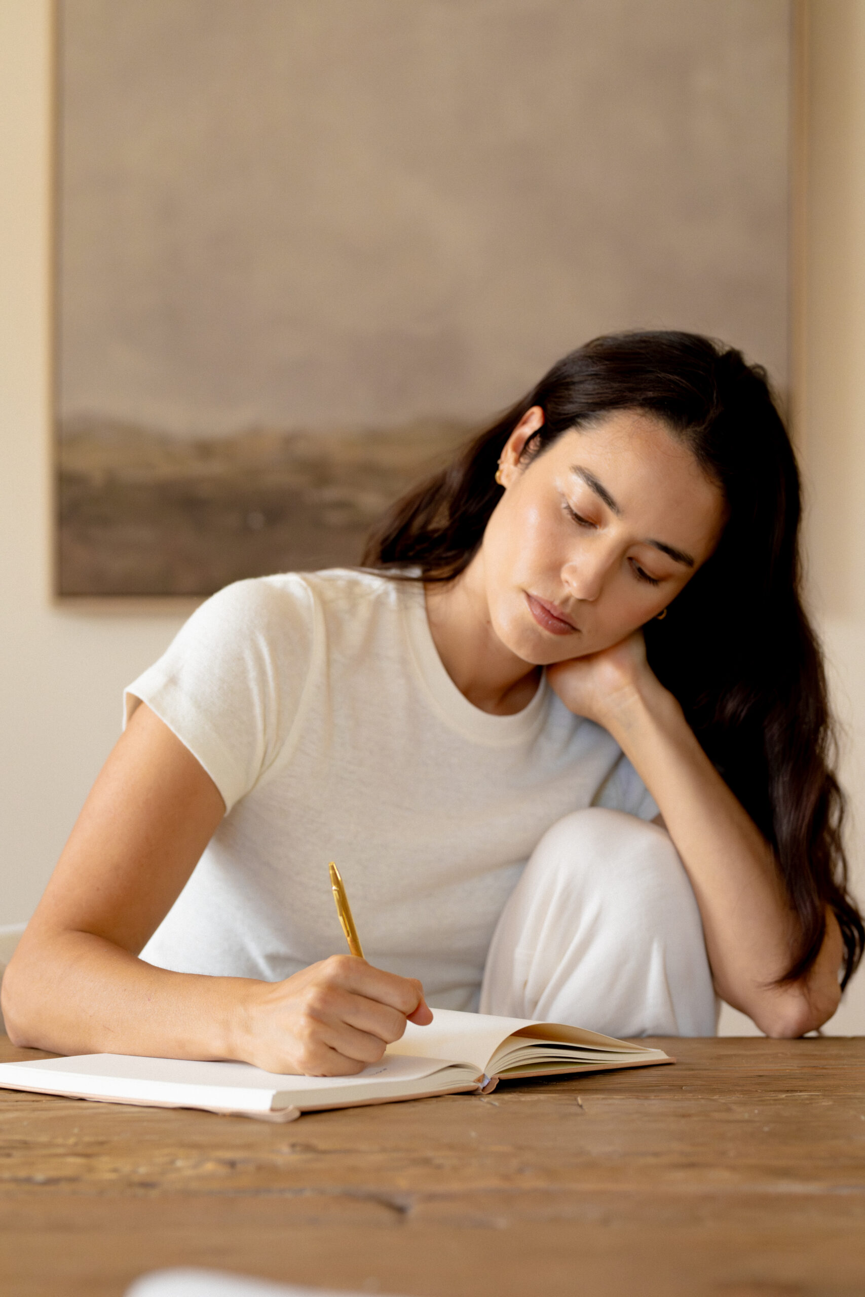 Woman in a white shirt sits at a wooden table, writing in a notebook with a pen, resting her head on her hand.