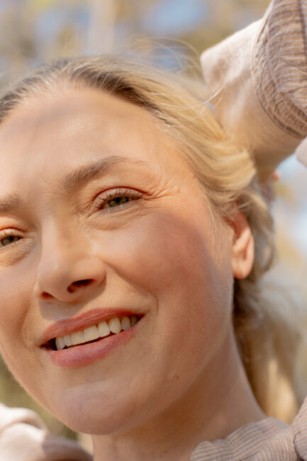 Close-up of a woman with light skin and blonde hair smiling outdoors, with sunlight and shadows on her face.
