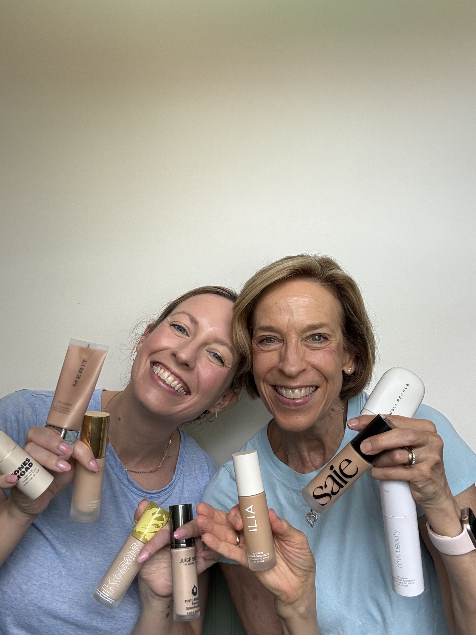 Two women smiling and holding various makeup foundation bottles in front of a plain white background.