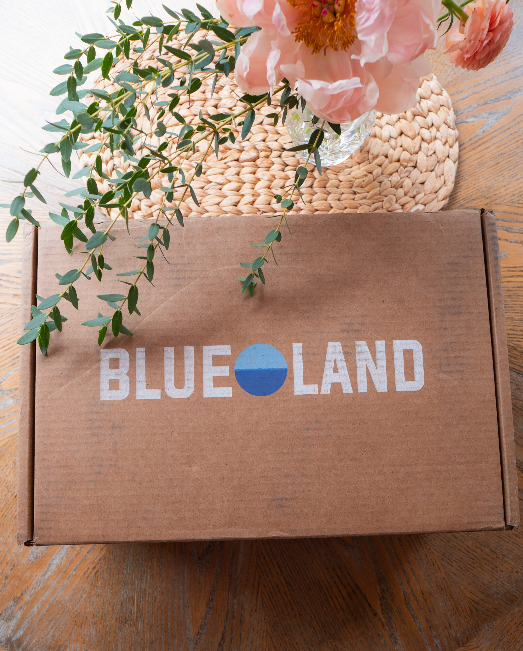 A Blueland-branded cardboard box sits on a wooden table next to a woven placemat and a vase with pink flowers and green leaves.