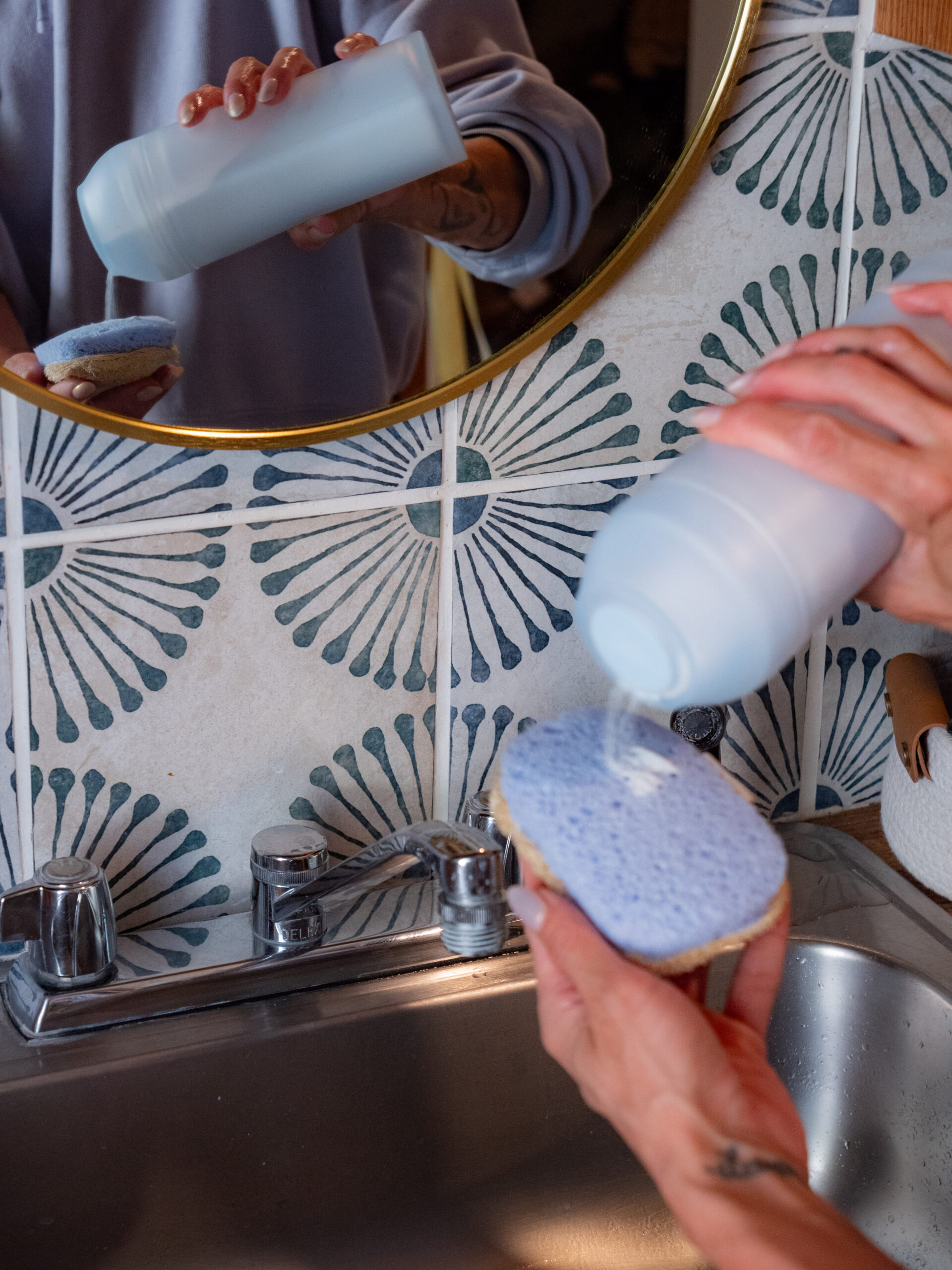 A person pours cleaning powder from a bottle onto a blue sponge over a kitchen sink, with a decorative tiled backsplash and a round mirror visible.