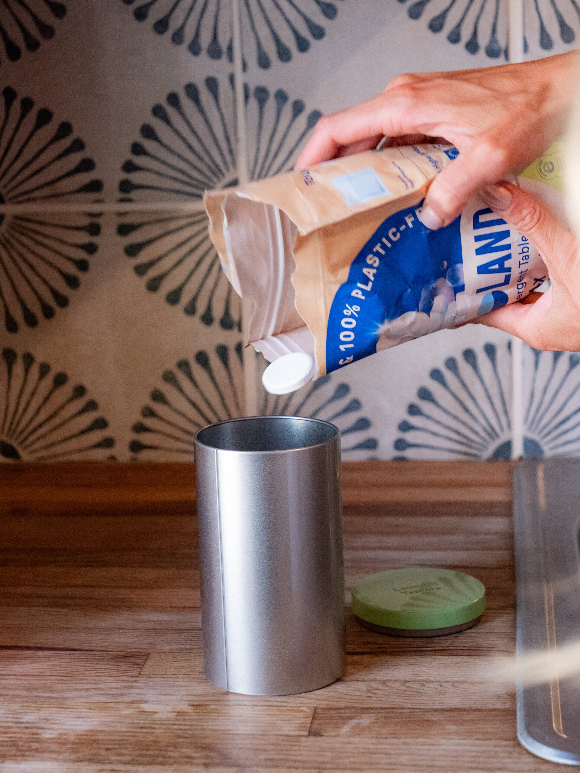 A person pours white dishwasher tablets from a paper bag into a silver storage container on a wooden counter.