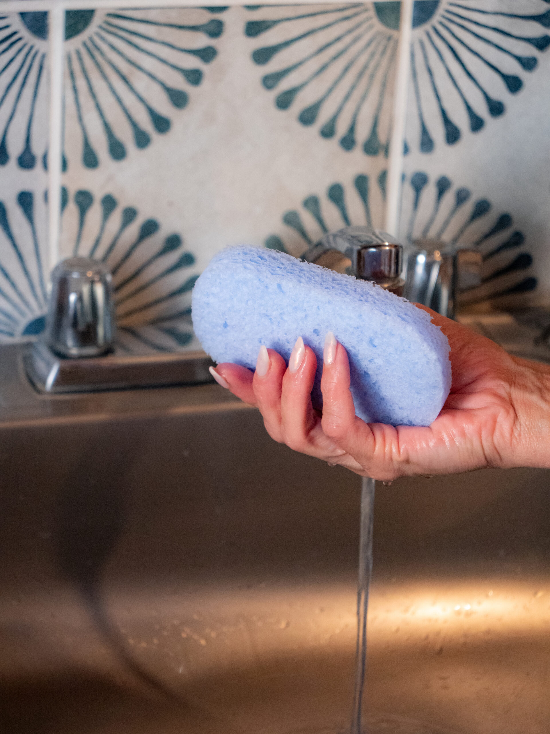 A hand holding a blue sponge under running water from a sink faucet, with patterned tiles in the background.