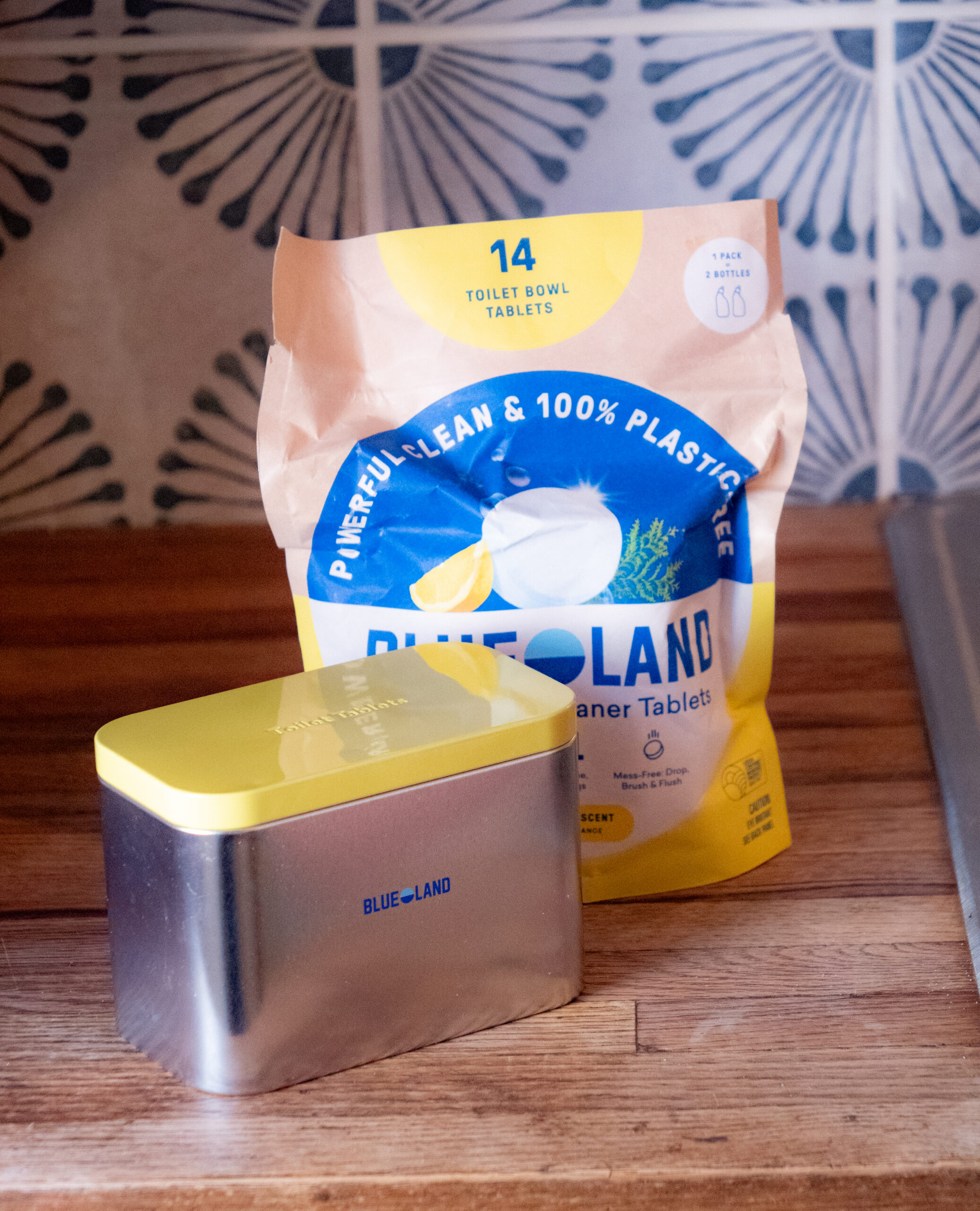 A pack of Blueland toilet bowl cleaner tablets and a metal storage tin with a yellow lid are on a wooden countertop.