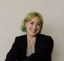 A person with short green hair, gold hoop earrings, and visible tattoos on their chest, wearing a black blazer, smiles at the camera against a plain background.