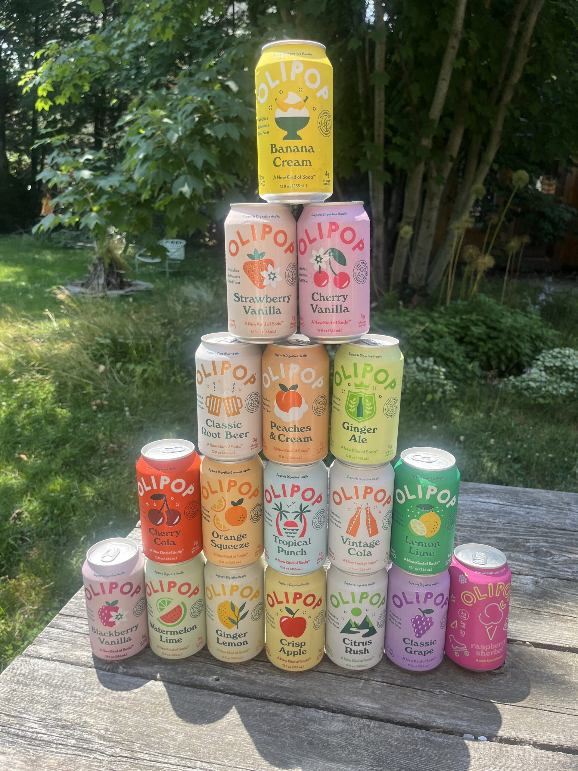 A pyramid of sixteen colorful OLIPOP soda cans in various flavors is displayed on a wooden table outdoors, with trees and greenery in the background.