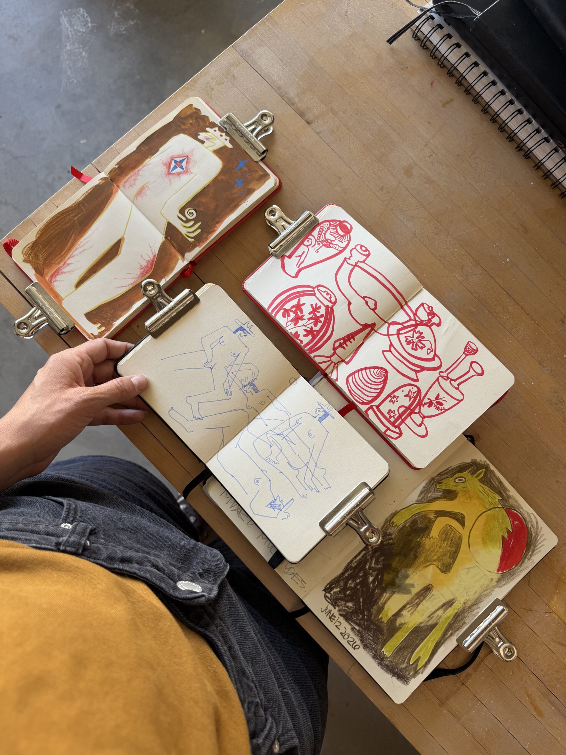 A person holds four sketchbooks open on a table, displaying colorful drawings and sketches, with metal clips holding the pages flat.