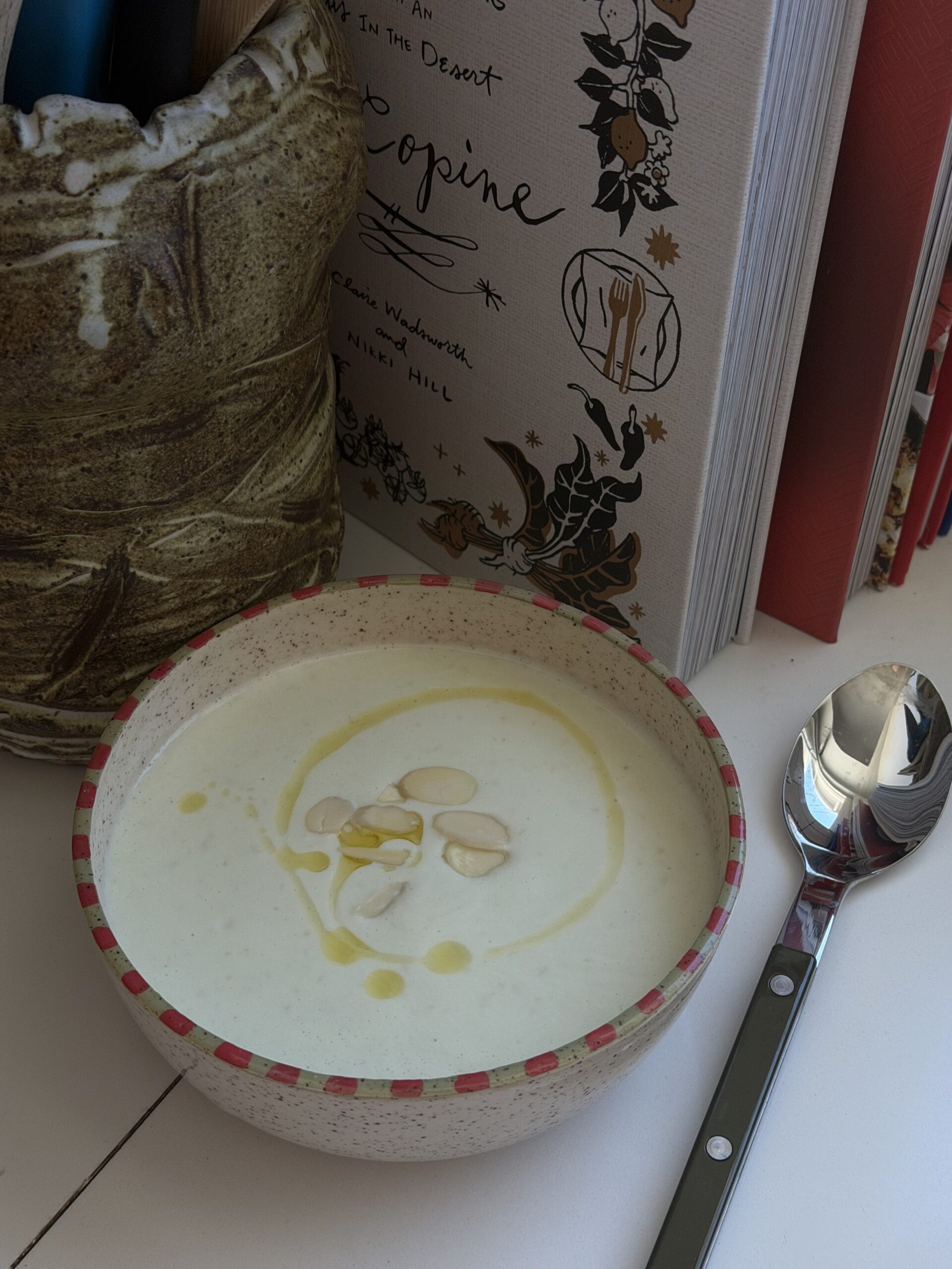 A bowl of creamy white soup garnished with almond slices and a drizzle of oil sits on a white surface next to a spoon, a ceramic container, and a cookbook.