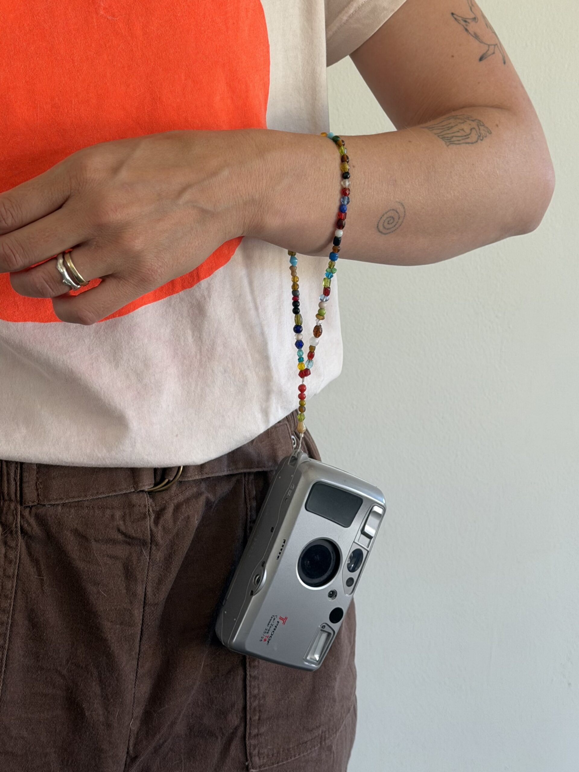 Person wearing a white and red shirt and brown pants holds a silver film camera with a beaded strap. Visible arm tattoos and two rings on their hand.