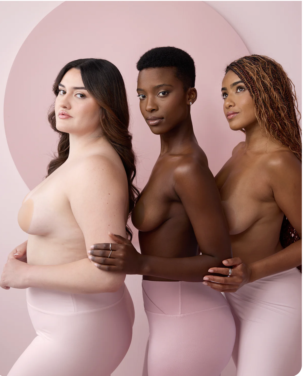 Three topless women of different skin tones pose side by side against a pink background, wearing pale pink pants and facing the camera with neutral expressions.