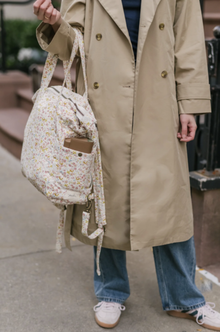 Person wearing a beige trench coat and blue jeans holding a floral patterned bag on a city sidewalk.