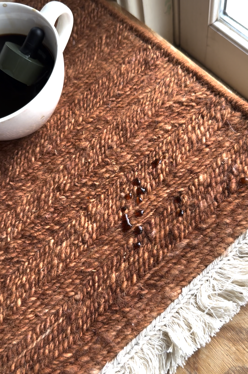 A white cup of dark liquid is tipped on a brown woven rug, causing several small dark stains on the rug near the cup.