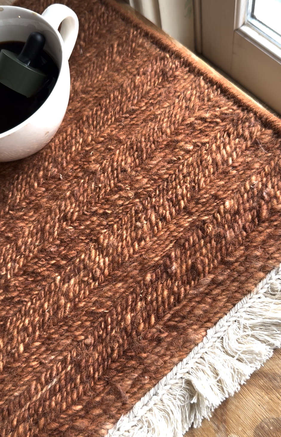 A white mug with dark liquid sits on a textured brown woven mat with fringe, placed beside a window.