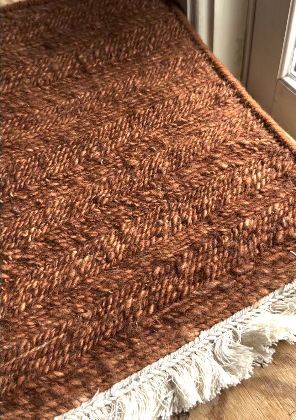 A close-up of a brown textured woven rug with a herringbone pattern and white fringe on one edge, positioned next to a glass door.