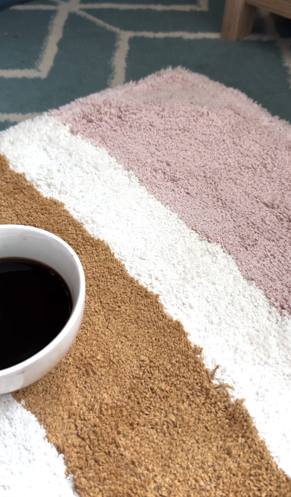 A white cup filled with black coffee is placed on a striped rug with brown, white, and pink sections, next to a blue and white patterned carpet.