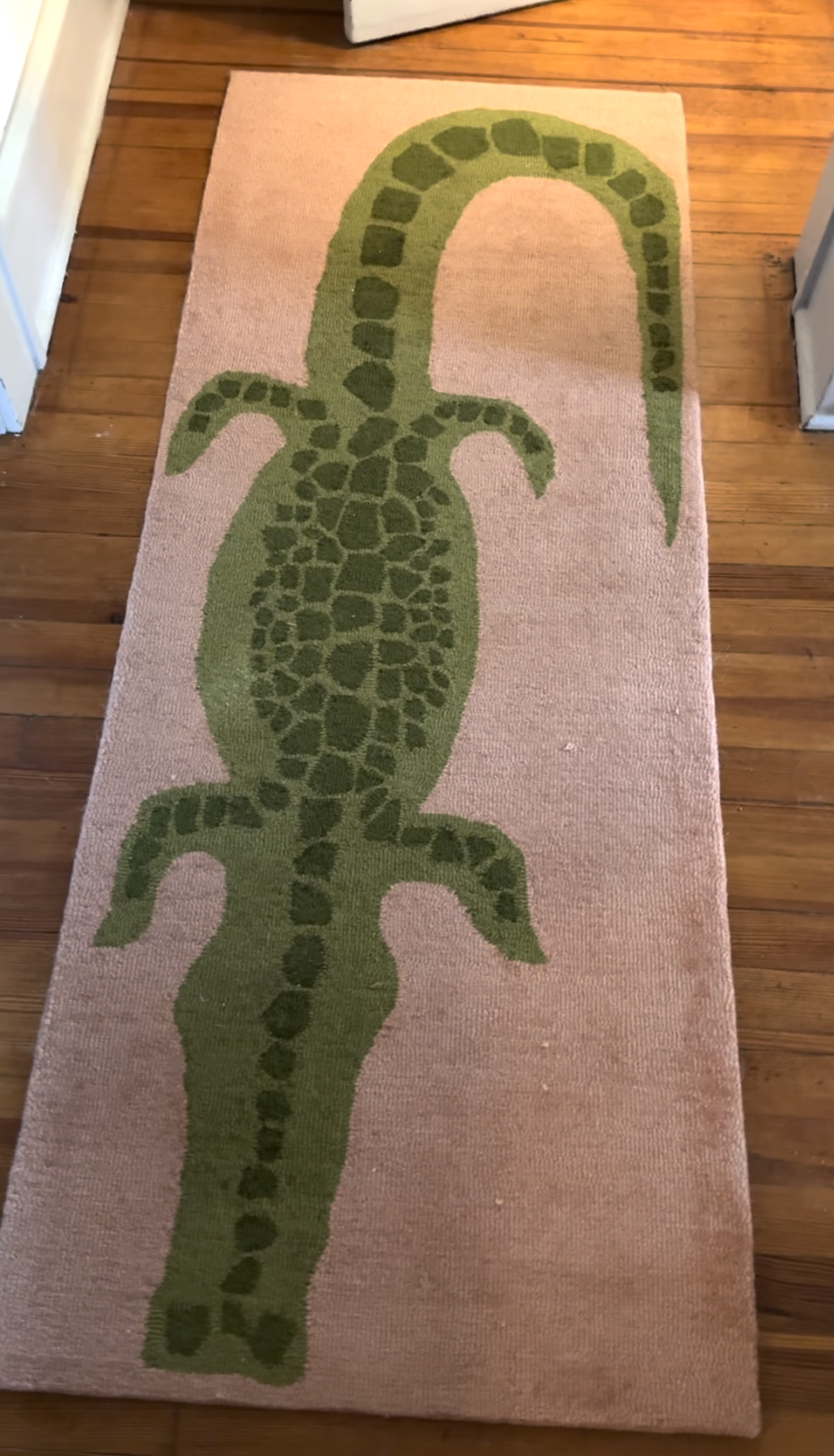 Rectangular rug with a green crocodile design on a light brown background, placed on a wooden floor between two doorways.