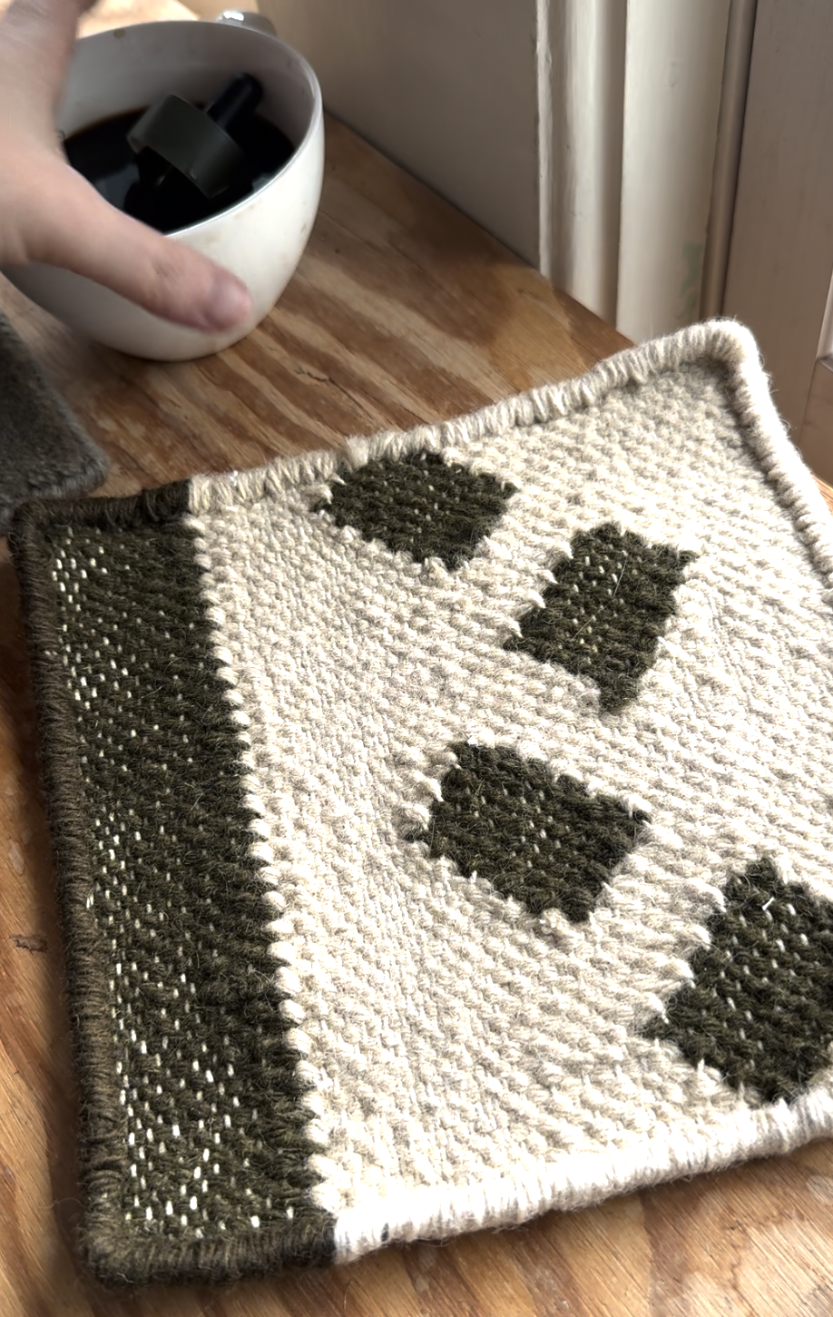 A hand holds a white cup near a woven, rectangular, cream and olive green placemat with geometric patterns, placed on a wooden surface.
