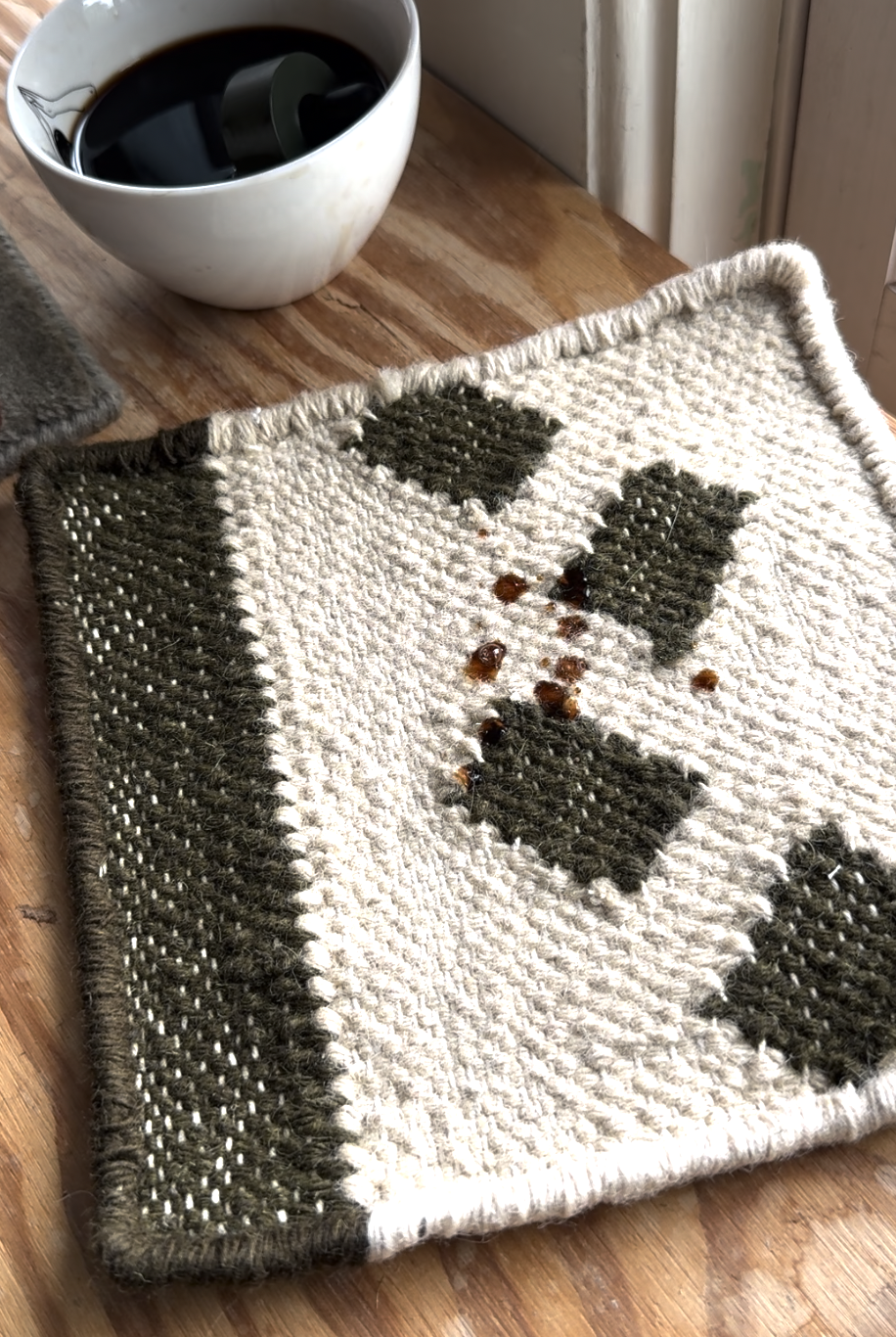 A white and green woven coaster with spilled coffee drops on it sits on a wooden table next to a white cup of black coffee.