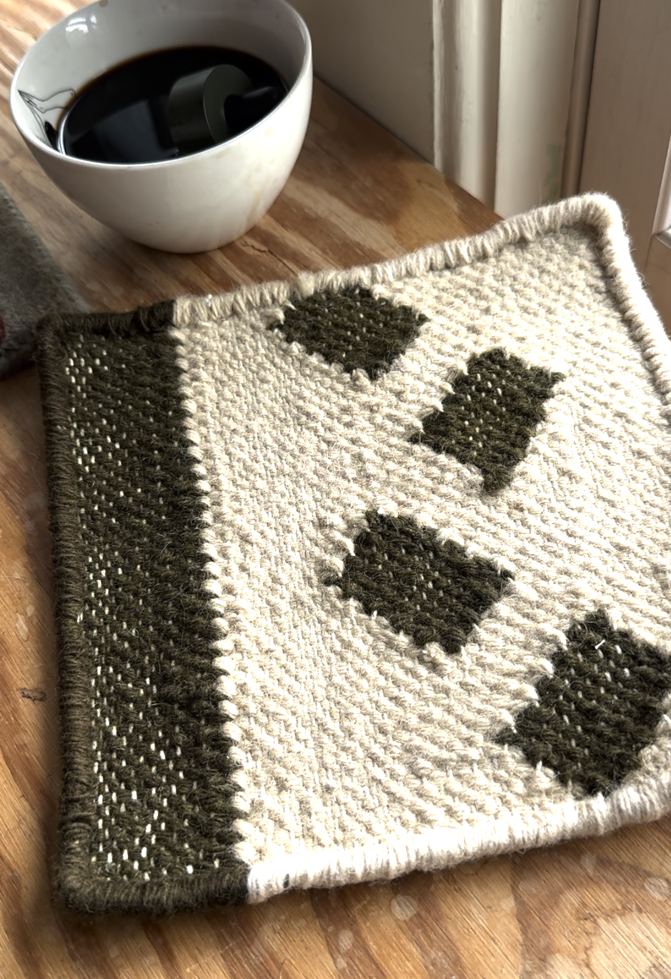 A woven coaster with green and cream geometric patterns sits on a wooden surface near a white cup filled with a dark liquid.