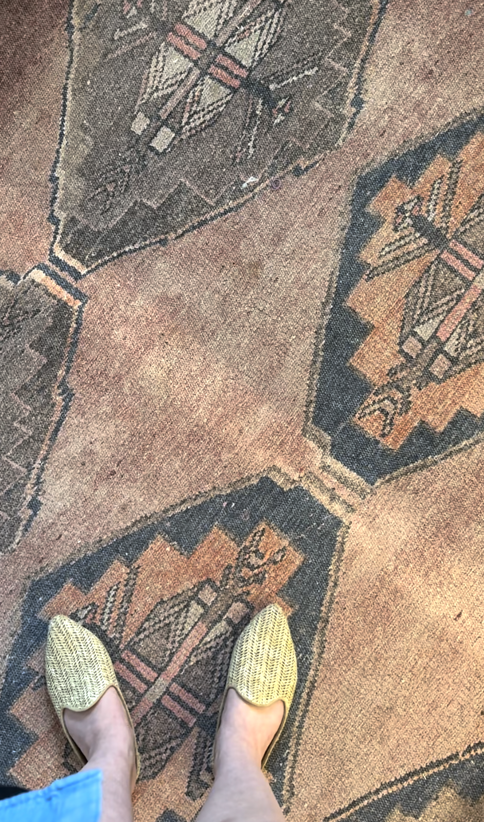 Person wearing woven shoes standing on a patterned rug with geometric designs and earthy tones.