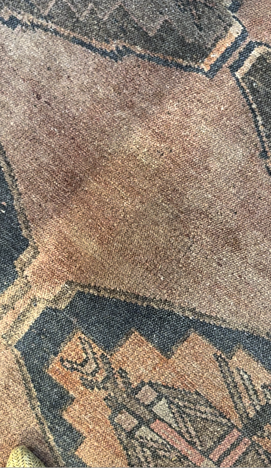 Close-up of a worn, patterned rug with faded earth-tone colors and geometric shapes, showing visible texture and signs of use.