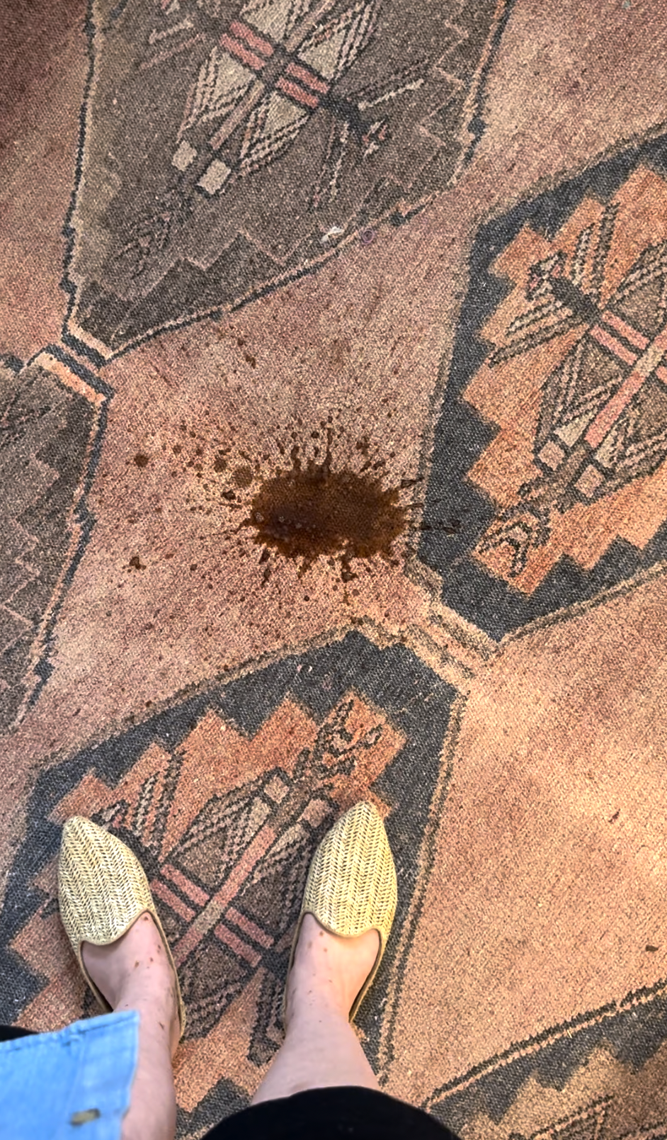 A person wearing woven shoes stands on a patterned carpet with a fresh brown liquid spill in the center of the image.