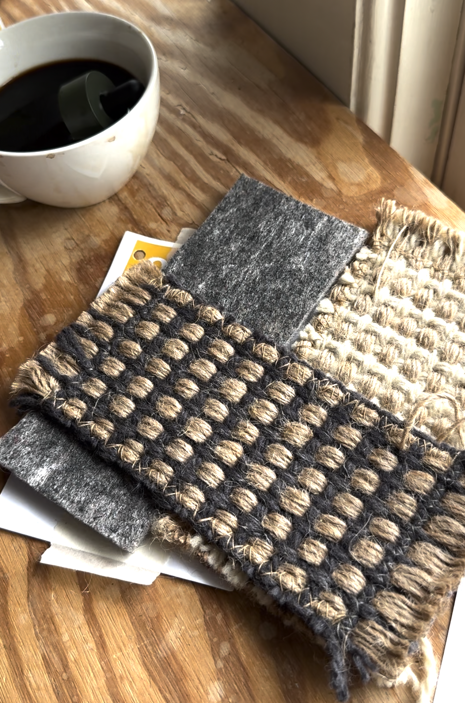 A cup of coffee sits next to several woven and felted textile samples on a wooden table near a window.