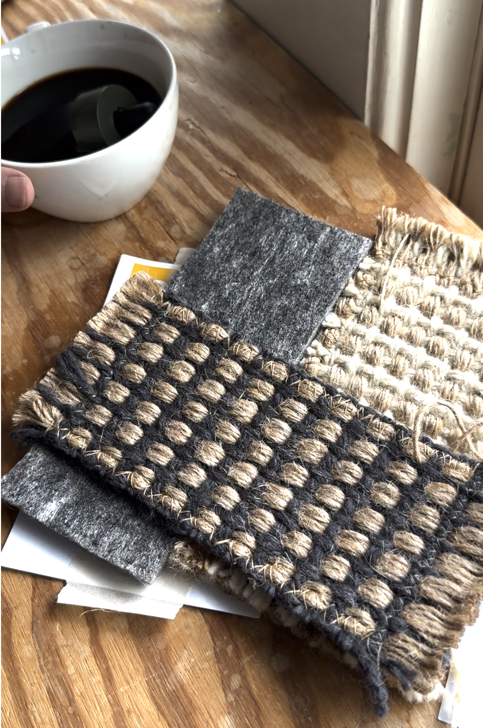 A hand holds a cup of coffee next to a wooden table with several textured fabric and carpet swatches in neutral tones spread out.