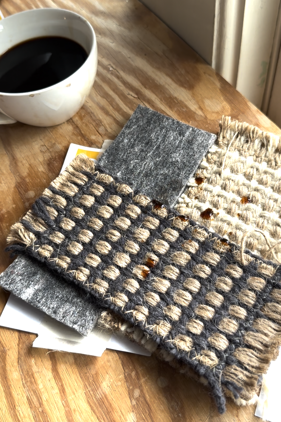 A cup of coffee sits on a wooden table next to several stacked fabric and textile swatches in gray, beige, and woven patterns.