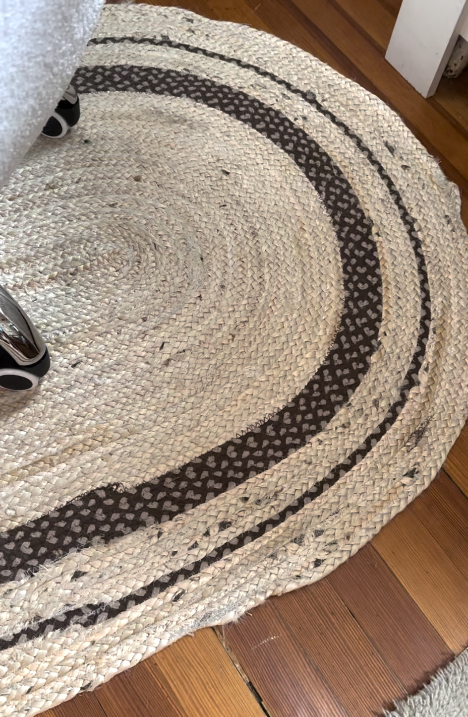 Oval braided rug with dark-striped pattern on a wooden floor, partially under a desk chair with wheels.