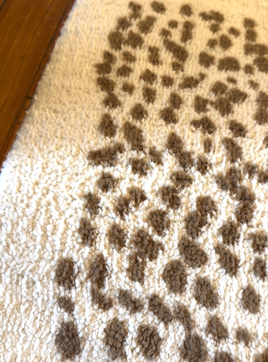 Close-up of a white textured rug with irregular brown spotted patterns, next to a wooden floor.