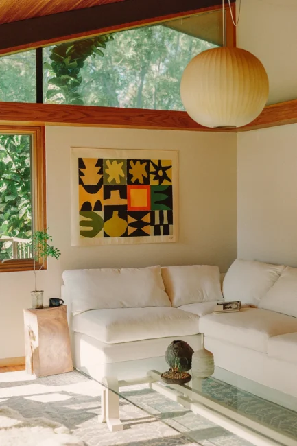 A modern living room with a white sectional sofa, abstract art on the wall, a glass coffee table, and a large round ceiling light. Sunlight streams through large windows.