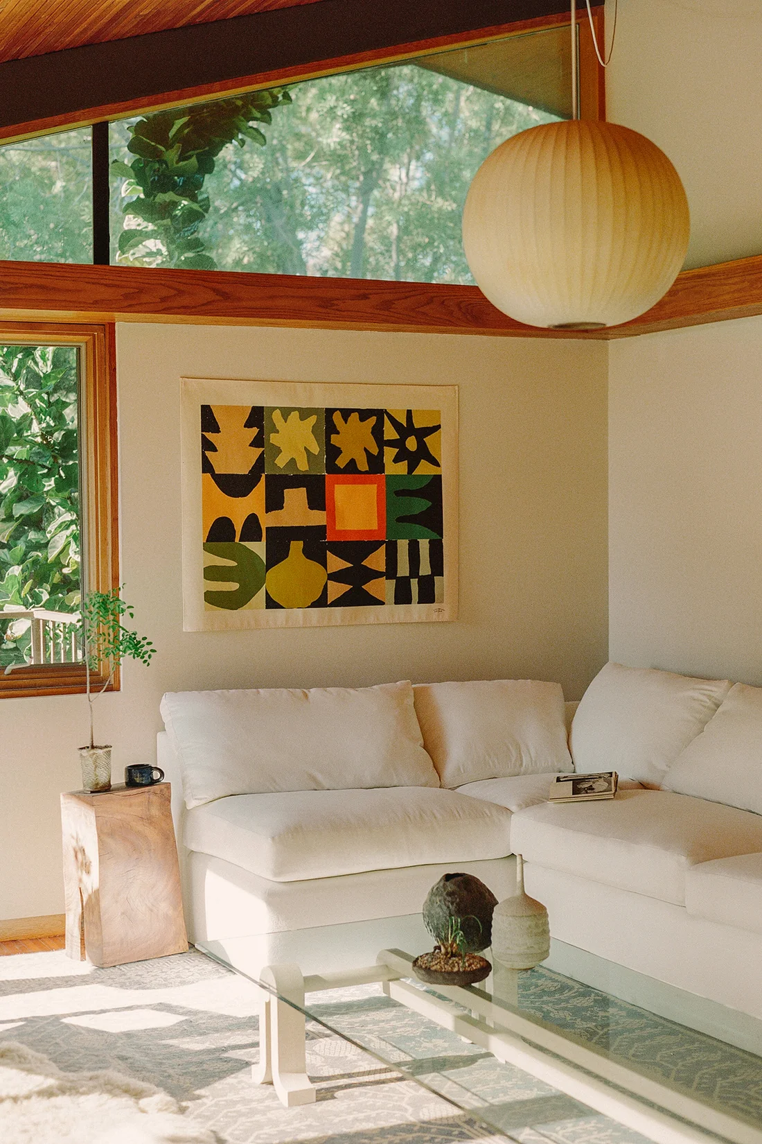 A modern living room with a white sectional sofa, abstract art on the wall, a glass coffee table, and a large round ceiling light. Sunlight streams through large windows.
