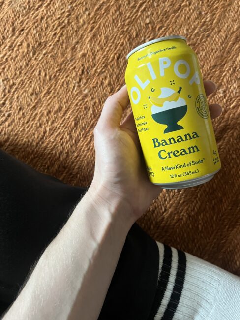 A hand holds a yellow can of OLIPOP Banana Cream soda over an orange textured surface, with part of a black pant leg and a white sock visible.
