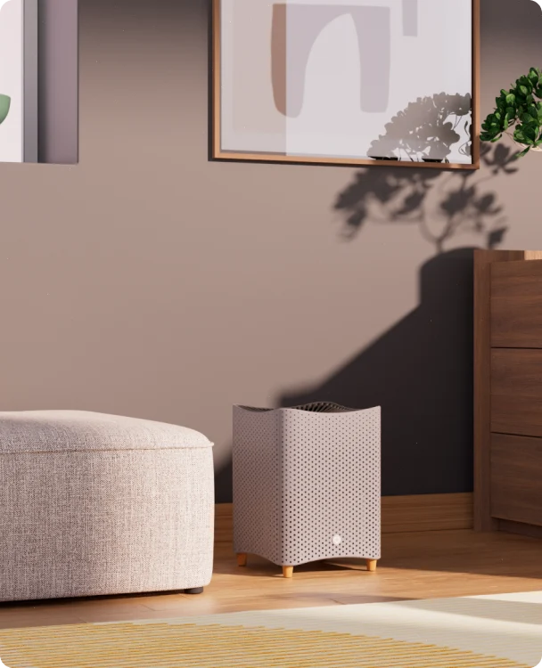 A small, gray air purifier sits on a wooden floor next to a beige ottoman and a wooden dresser, with framed art and plant shadows on the taupe wall behind.