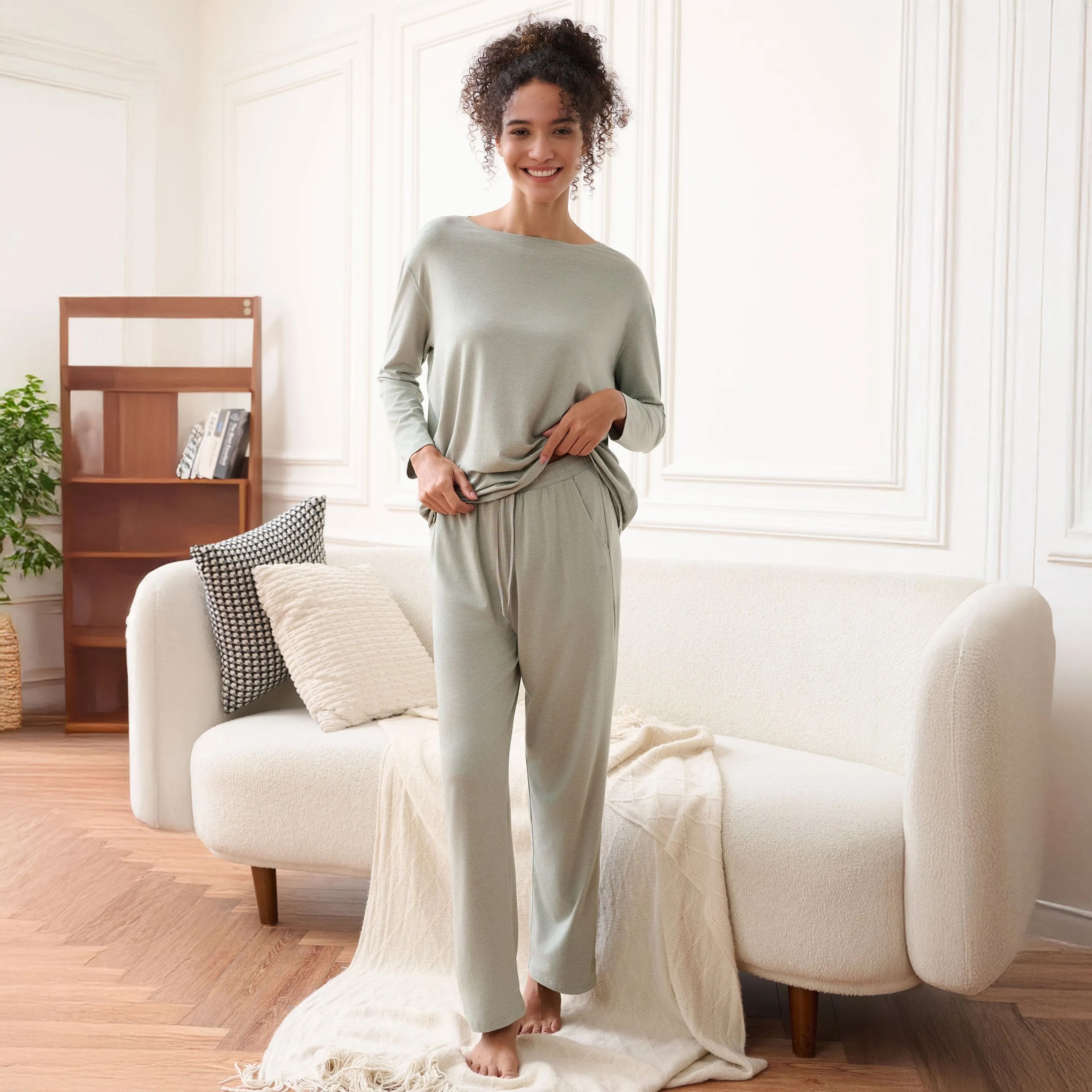 Woman standing barefoot in a living room, wearing light gray loungewear and smiling, in front of a cream sofa with pillows and a blanket, with a wooden shelf and plant in the background.