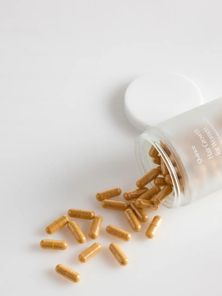 A clear bottle labeled “Hair Growth for Women” lies on its side with brown capsules spilling out onto a white surface.
